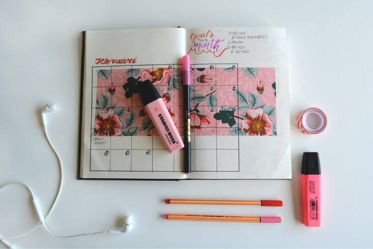 Caderno de anotações com calendário desenhado. Ao redor, canetas coloridas.