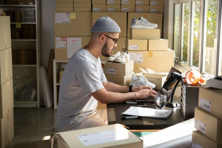 Homem no notebook. Ao redor, caixas de produtos de seu e-commerce.