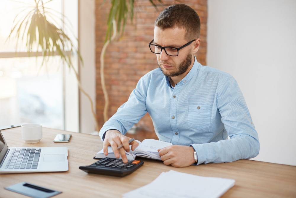 Homem calcula finanças com calculadora e anotações, representando a análise da margem de lucro em marketplaces.