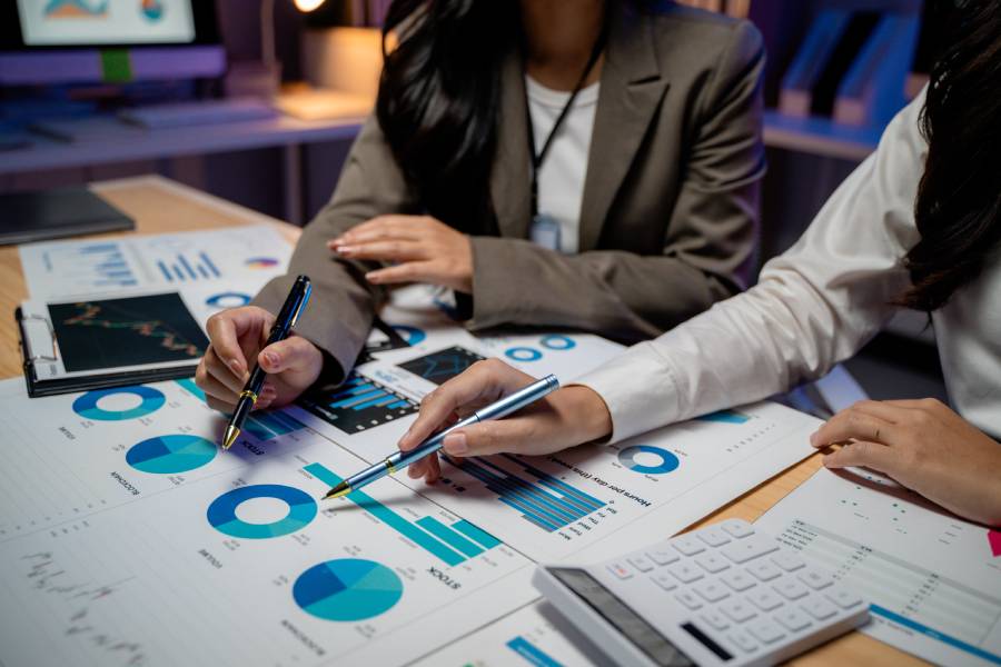 Duas pessoas apontando canetas sobre relatórios com gráficos em azul espalhados em uma mesa, analisando resultados de campanhas da Amazon