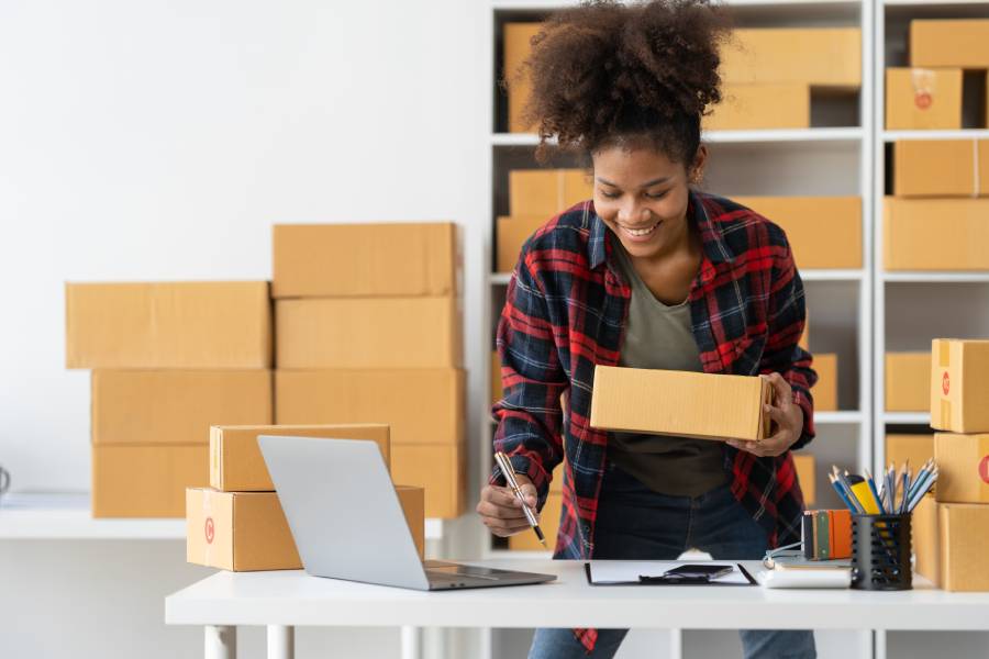 Mulher sorridente segurando caixa de papelão e anotando dados em laptop, gerenciando crescimento em marketplace