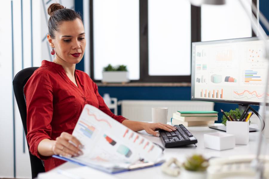 Profissional realizando análise de DRE com gráficos em monitor e documentos financeiros sobre a mesa