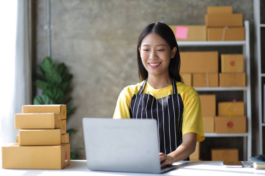 Vendedora com avental usando laptop em ambiente com caixas, fazendo a conciliação do Mercado Livre
